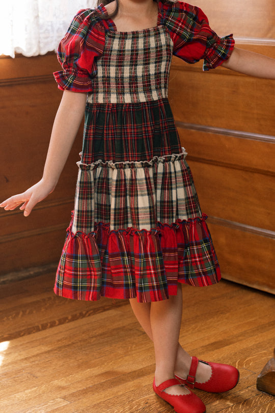 A child wearing the Mini Madeline Dress in Mixed Plaid by Ivy City Co—a delightful girls’ holiday dress with puffed sleeves and ruffle details in classic red and green plaid—stands on a wooden floor in a warmly lit room, her red shoes finishing the festive look for a perfect Christmas ensemble.