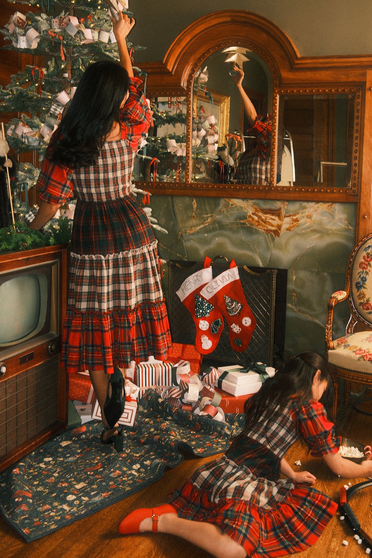 Two girls in matching Mini Madeline Dress in Mixed Plaid by Ivy City Co decorate a Christmas tree. One stands reaching up, while the other kneels on the floor arranging gifts. A vintage TV, stockings, and wrapped presents sit near a decorated mirror.