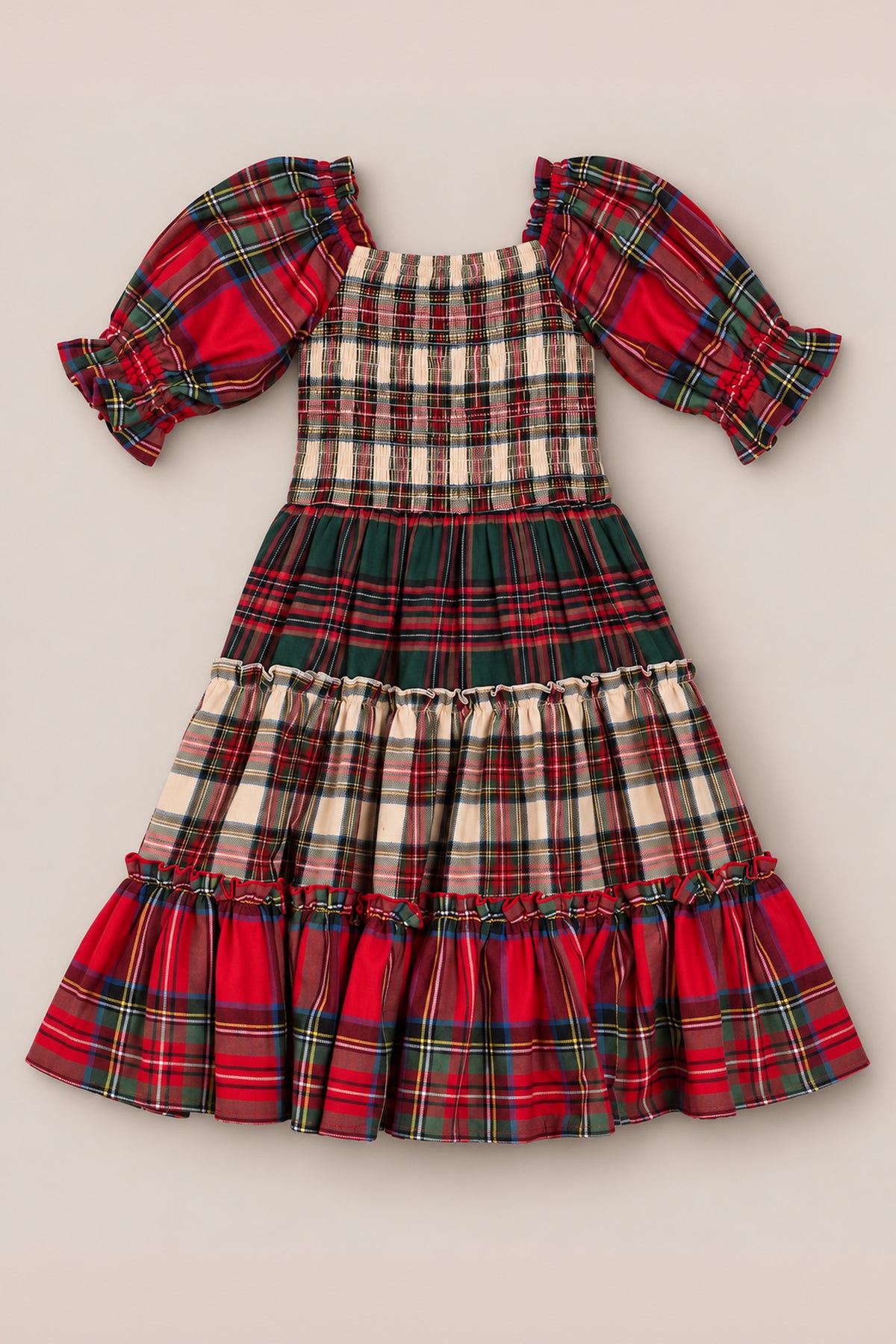 The Mini Madeline Dress in Mixed Plaid by Ivy City Co is a delightful girls’ plaid dress with puff sleeves and a ruffled hem, showcasing festive red, green, and cream patchwork patterns on a neutral background—making it the perfect Christmas dress for the holiday season.