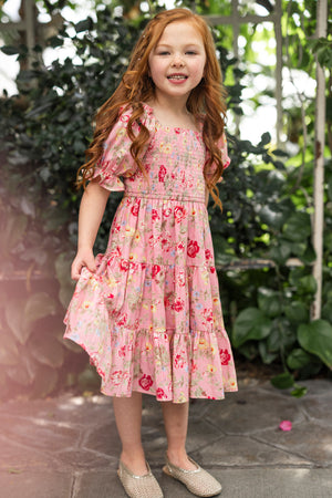 A young girl with long red hair wears the Ivy City Co Mini Madeline Dress in Pink Vintage Poppies and sparkly shoes, smiling as she holds her dress while standing on a stone path surrounded by lush green plants.