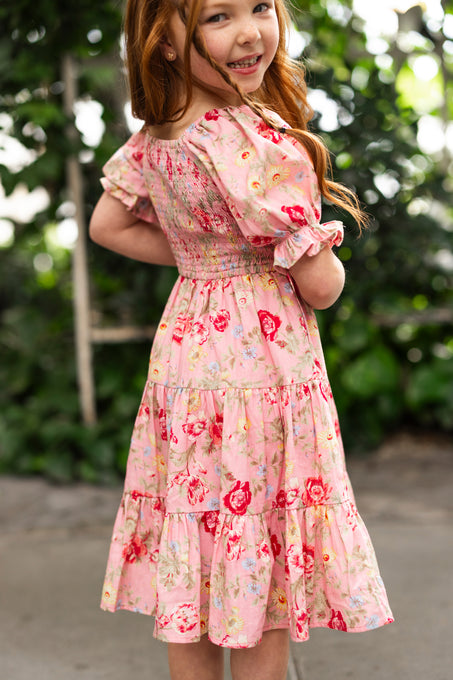A young girl with long red hair smiles while wearing the Ivy City Co Mini Madeline Dress in Pink Vintage Poppies, a charming spring dress. She stands outdoors amid greenery and blurred foliage, turned slightly to the side.