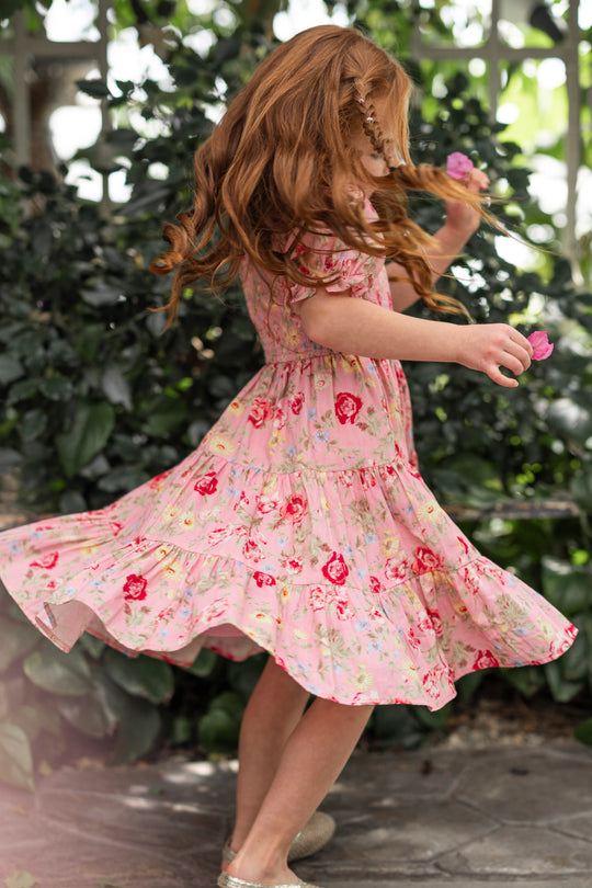 A young girl with long, wavy red hair twirls joyfully outdoors in Ivy City Co’s Mini Madeline Dress in Pink Vintage Poppies, surrounded by lush green plants and stone pavement.