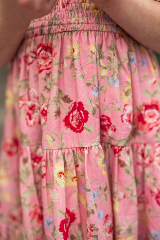 Close-up of the Mini Madeline Dress in Pink Vintage Poppies by Ivy City Co, a girls’ spring dress with red, yellow, and blue flowers plus green leaves, gathered tiered fabric, and part of an arm visible at the image edge.