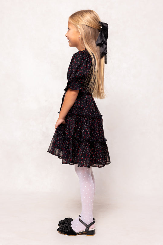 Young girl wearing a black floral dress with white tights and black shoes on a plain background