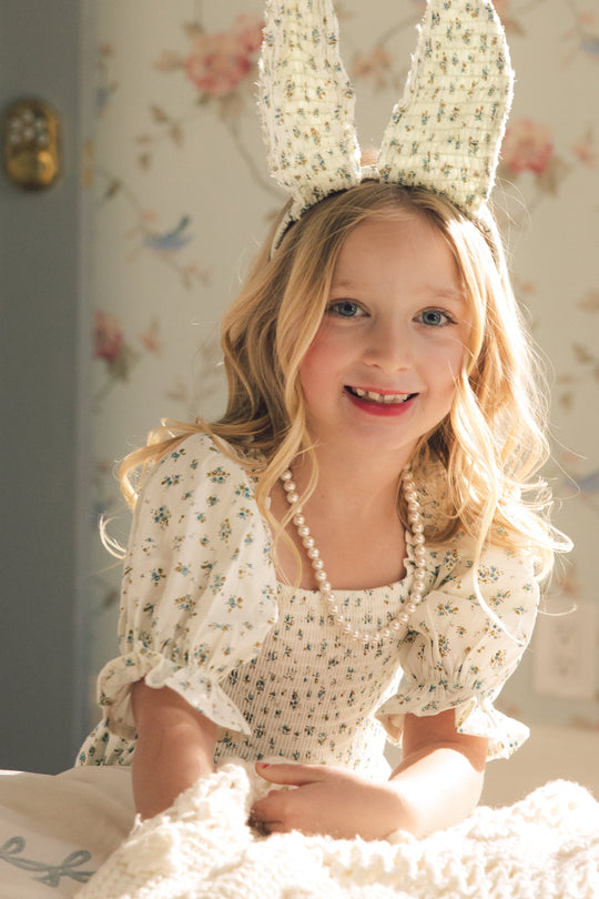 A smiling young girl with long blonde hair wears a bunny ear headband and the Mini Madeline Dress in Blue by Ivy City Co, featuring a smocked bodice and pearl necklace, as she sits indoors with soft sunlight and floral wallpaper in the background.