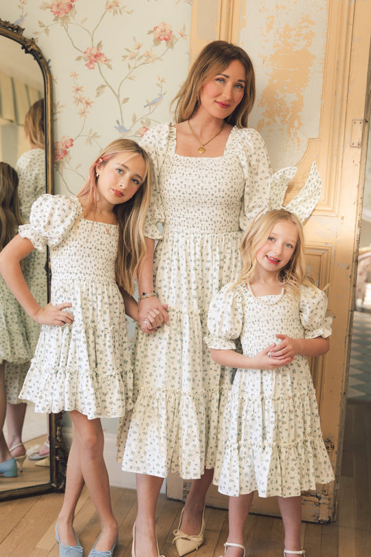 A woman and two young girls stand together indoors wearing matching Ivy City Co Mini Madeline Dresses in Blue, each featuring a smocked bodice, posing and holding hands, with a tall mirror and floral wallpaper in the background.