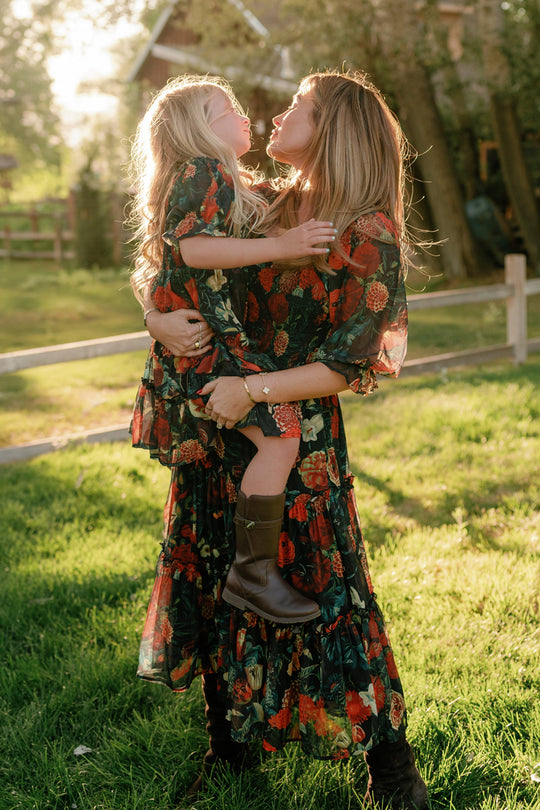 A woman and a young girl, both wearing the Ivy City Co Mini Madeline Dress in Imogen Moody Floral, stand outside in the sunlight. The dresses feature airy chiffon fabric and smocked bodices. The woman holds the girl as they smile at each other in a grassy yard surrounded by trees and a wooden fence.