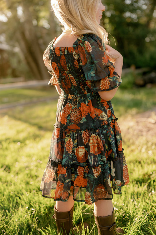 A child with long blonde hair, wearing the Ivy City Co Mini Madeline Dress in Imogen Moody Floral—crafted from airy chiffon with a smocked bodice—stands on grass in warm sunlight, facing away from the camera.