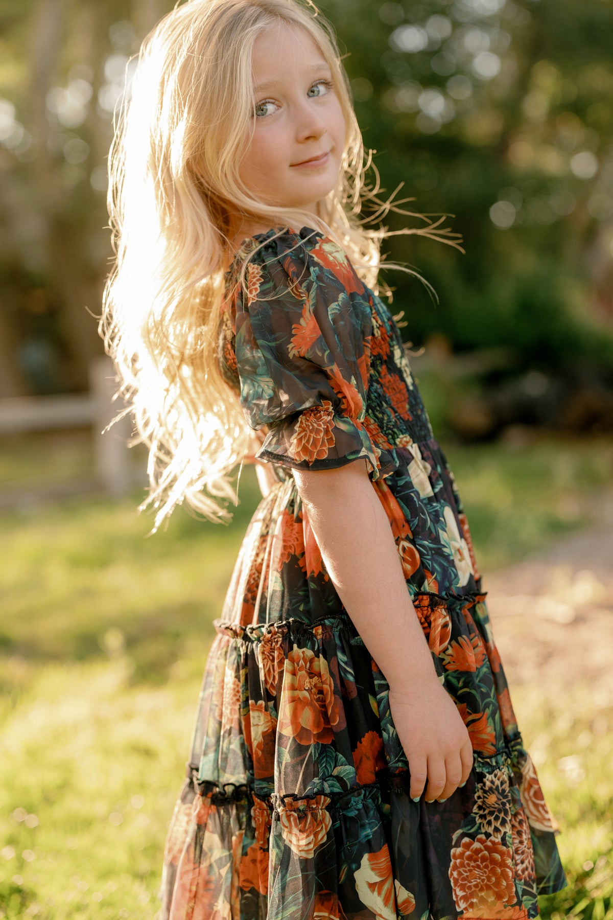 A young girl with long blonde hair stands outdoors in sunlight, wearing the Mini Madeline Dress in Imogen Moody Floral by Ivy City Co— a dark chiffon dress adorned with orange and green floral patterns. She looks over her shoulder with a gentle expression, set against a backdrop of blurred greenery.