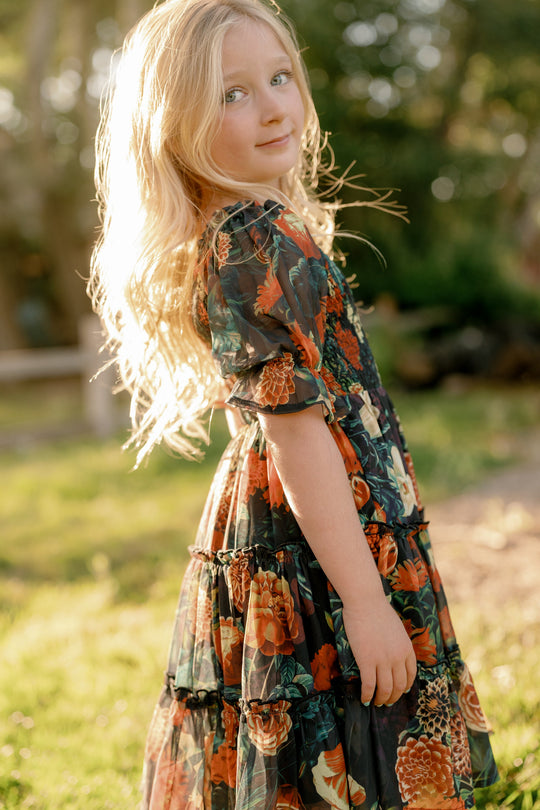 A young girl with long blonde hair stands outdoors in sunlight, wearing the Ivy City Co Mini Madeline Dress in Imogen Moody Floral and looking back over her shoulder with a gentle expression.