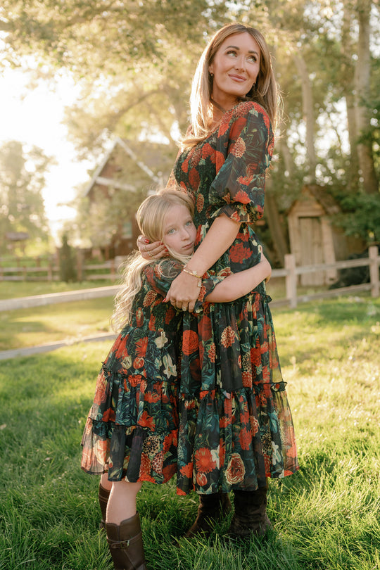 A woman and a young girl embrace on the grass, both wearing the Ivy City Co Mini Madeline Dress in Imogen Moody Floral, as sunlight streams through the trees with a wooden fence and cabin in the background.