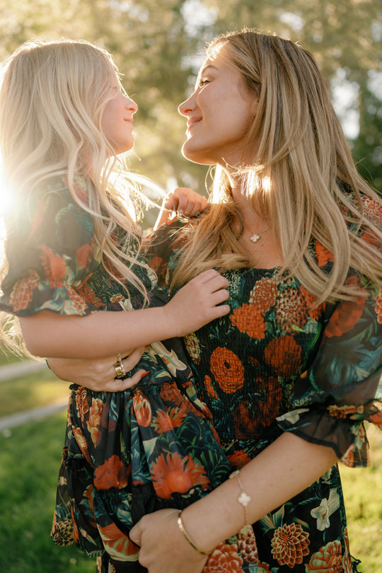 A woman and a young girl, both wearing the Ivy City Co Mini Madeline Dress in Imogen Moody Floral, smile at each other in warm sunlight outdoors, sharing a joyful, loving moment.