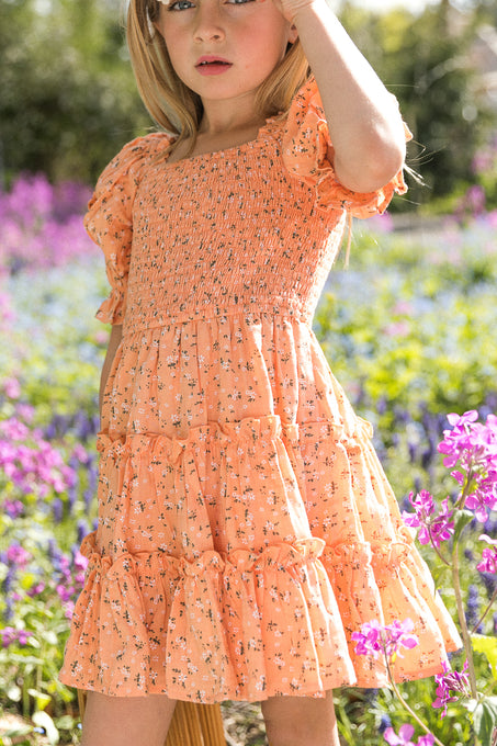 A young girl stands in a field of purple flowers wearing the Ivy City Co. Mini Madeline Dress in Sorbet, featuring a smocked bodice and puff sleeves, surrounded by lush greenery and blooms on a sunny day.