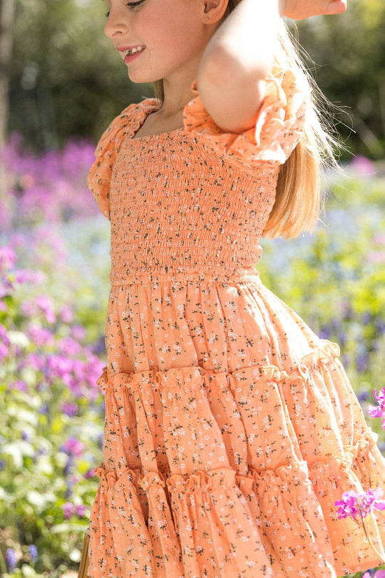 A young girl in the Ivy City Co. Mini Madeline Dress in Sorbet, featuring puff sleeves and a floral pattern, stands outdoors among purple and green wildflowers, smiling and enjoying a sunny day.