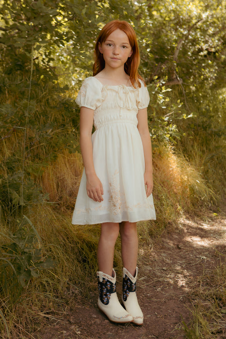 A young girl with red hair stands on a sunlit forest path, wearing Ivy City Cos Mini Mae Dress in Metallic Thread with short sleeves and hand-embroidered details, paired with white cowboy boots featuring dark designs.