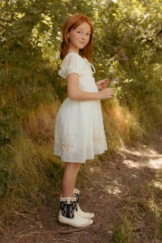 A young girl in the Ivy City Co Mini Mae Dress in Metallic Thread stands on a dirt path surrounded by green foliage, looking at the camera with a slight smile.