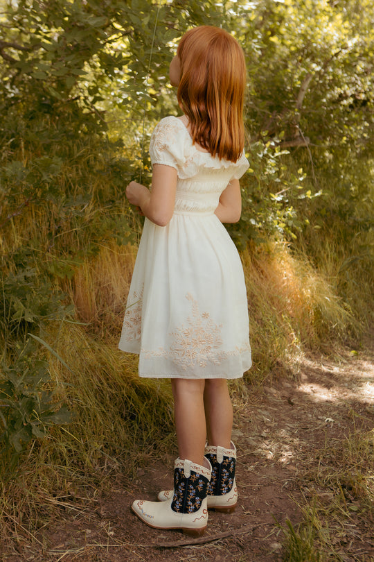 A young girl with red hair, wearing the Ivy City Co Mini Mae Dress in Metallic Thread and patterned cowboy boots, stands on a sunlit dirt path in a green forest, gazing up at the trees.