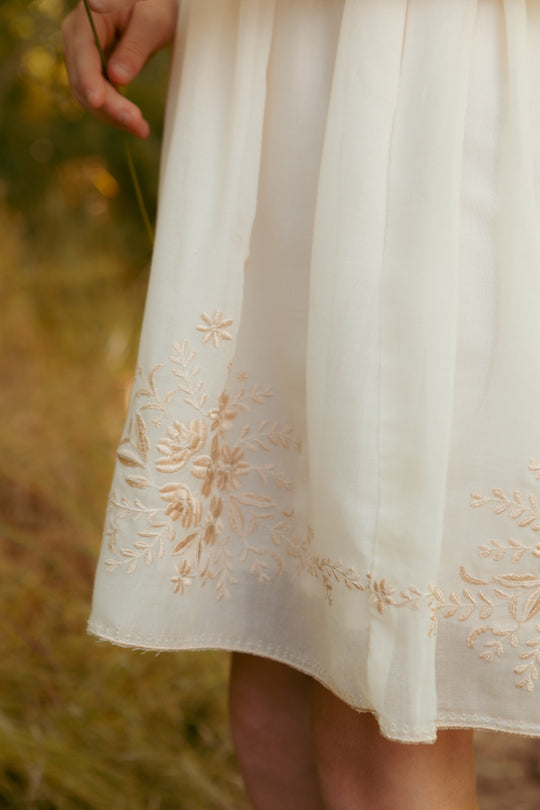 A close-up of a person wearing the Ivy City Co Mini Mae Dress in Metallic Thread, featuring delicate floral embroidery near the hem, standing outdoors in a grassy area. Only the lower dress and one hand are visible.
