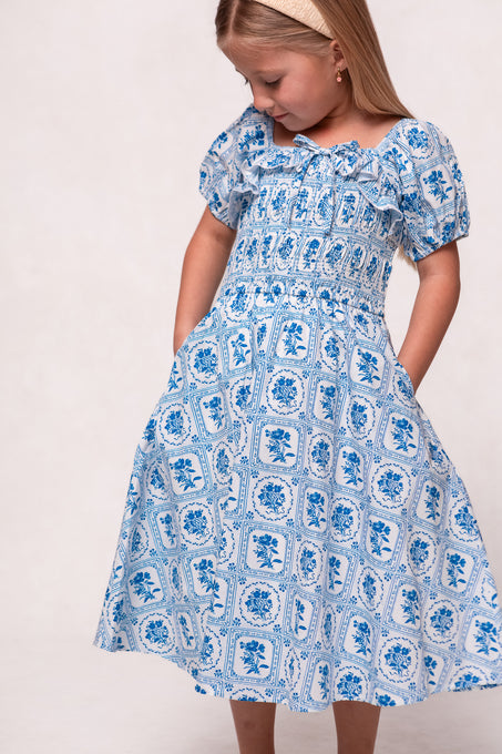 A young blonde girl in a beige headband and the Ivy City Co Mini Mae Dress in Blue Floral Stamp stands with her hands in the pockets, looking down and smiling softly.
