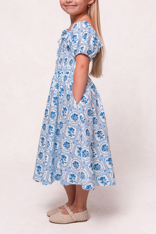 A young girl stands sideways in the Ivy City Co Mini Mae Dress in Blue Floral Stamp, featuring short sleeves and pockets, paired with beige woven shoes against a plain light background.