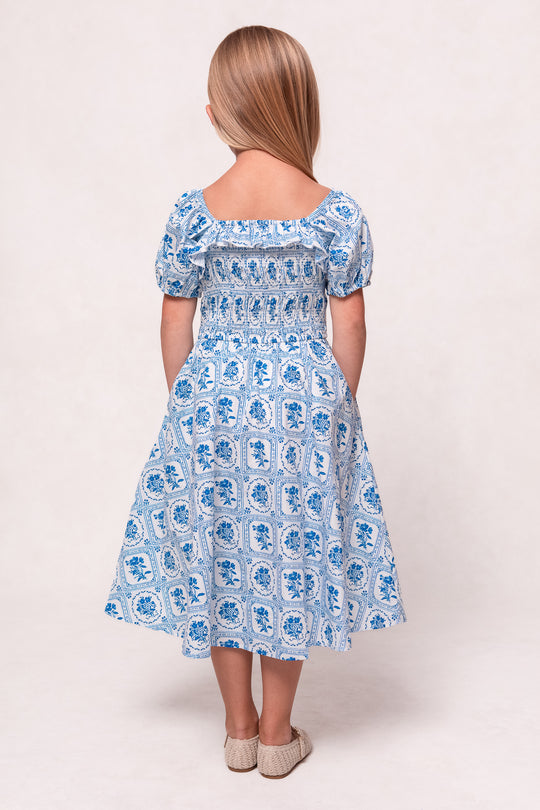 A young girl with straight, light brown hair stands facing away, wearing Ivy City Co’s Mini Mae Dress in Blue Floral Stamp and beige shoes, set against a light, neutral background.