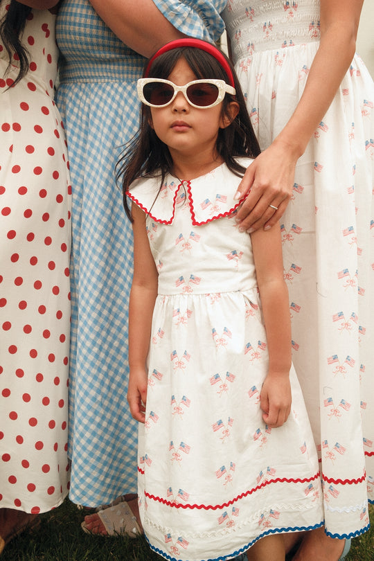 A young girl in white sunglasses and an Ivy City Co Mini Maisy Dress in Star Spangled stands before adults in vintage-inspired Fourth of July dresses, including red polka dots and blue gingham, as she looks ahead with an adult’s hand on her shoulder.