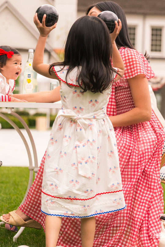 A girl in a white dress holds two black balls above her head as she faces a woman in red gingham. Nearby, a child sits at an outdoor table wearing the Ivy City Co Mini Maisy Dress in Star Spangled with a red bow.