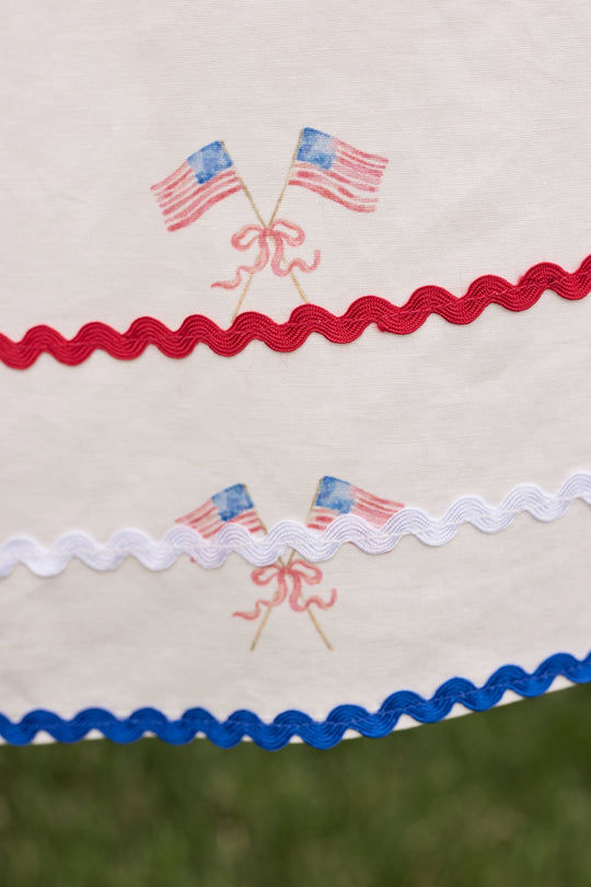 The Mini Maisy Dress in Star Spangled by Ivy City Co features white fabric with embroidered crossed American flags and wavy red, white, and blue trim—ideal for patriotic celebrations. The out-of-focus grassy background adds a playful touch.