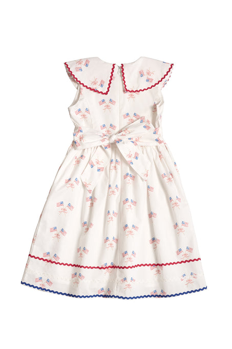 The Ivy City Co Mini Maisy Dress in Star Spangled is a white children’s dress with red and blue trim, a Peter Pan collar, waist bow, and American flag and fireworks print—perfect for the Fourth of July.