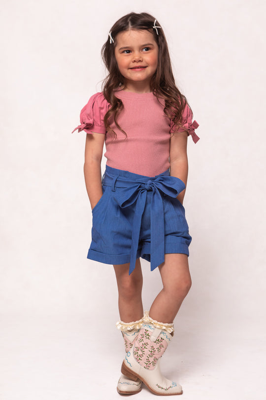 A young girl with long brown hair in pigtails wears a pink short-sleeve top, Ivy City Cos Mini Maria Shorts in Chambray with a tied belt, and white floral embroidered cowboy boots. She stands smiling with her hands in her pockets.