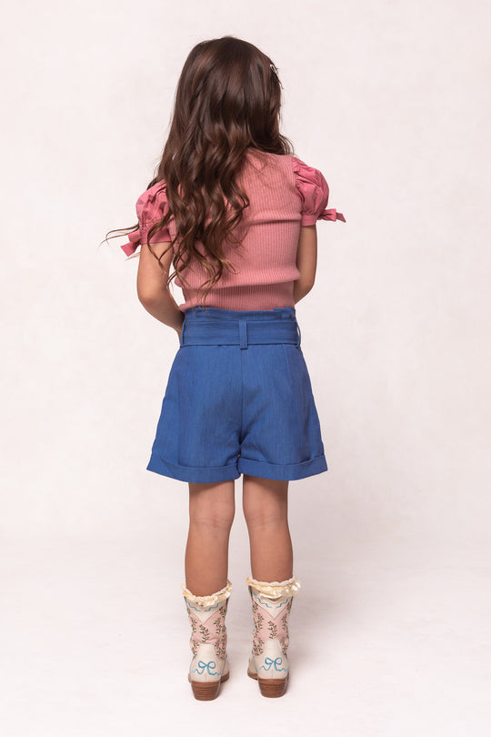 A young girl with long, wavy brown hair wears a pink short-sleeved top, Ivy City Co Mini Maria Shorts in Chambray, and patterned cowboy boots as she stands facing away from the camera against a light background.