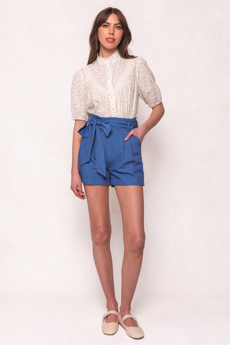 A woman with long brown hair wears a white, short-sleeved, high-collar button-up blouse and Ivy City Co’s Maria Shorts in Chambray with a tied belt. She stands against a light background, paired with beige flats.
