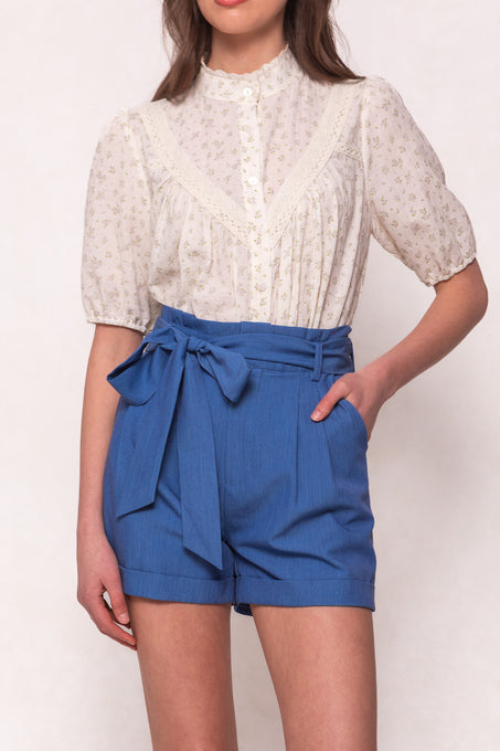 A woman wears the Ivy City Co Maria Shorts in Chambray, which have a high waist and tied belt, paired with a short-sleeve cream floral lace blouse. Her left hand is in her pocket, and her head is partially out of frame.