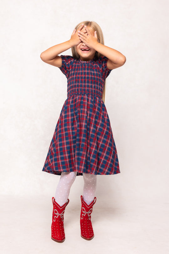 Child wearing a red and blue checkered dress with white tights and red shoes on a white background