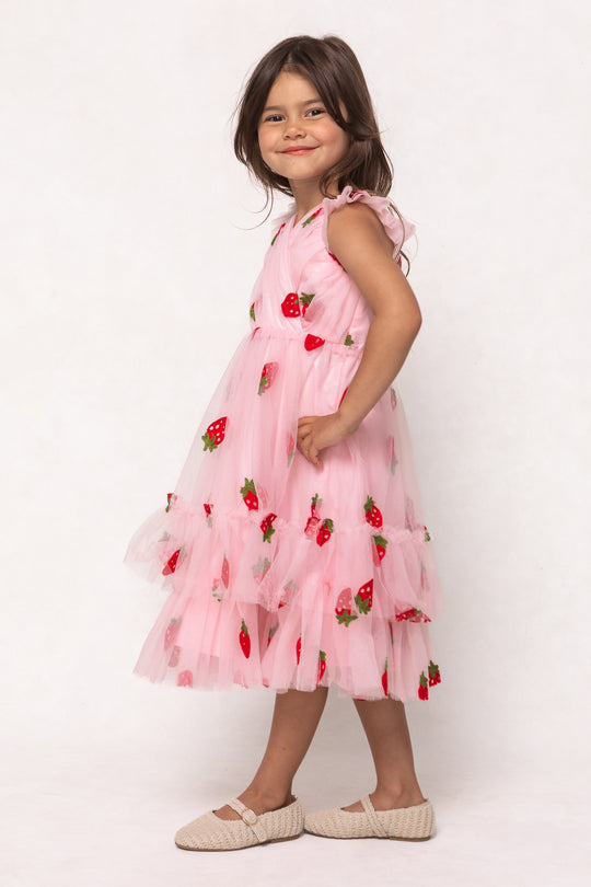 A young girl with long brown hair smiles in the Ivy City Co Mini Marin Dress in Strawberry Shortcake, a pink tulle dress with strawberry embroidery, paired with beige woven shoes as she stands sideways against a light background.
