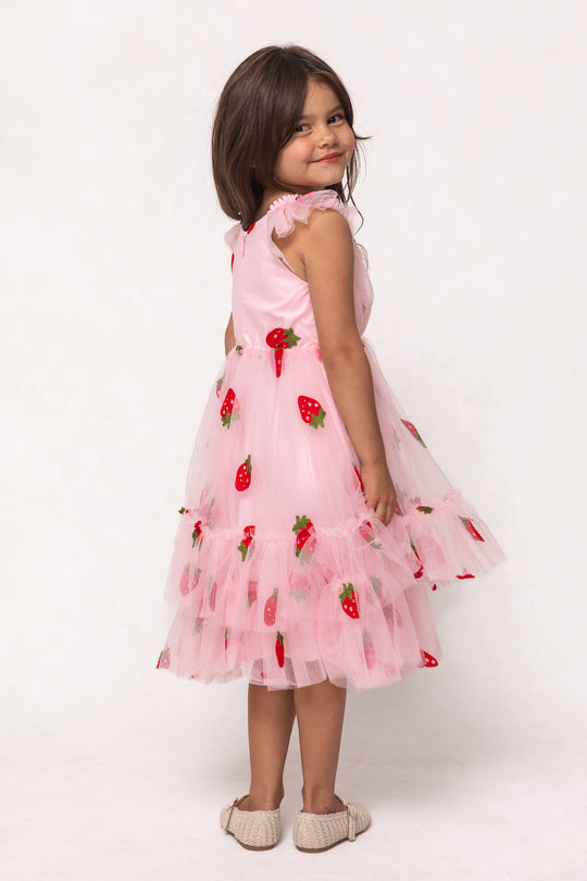 A young girl with brown hair smiles while wearing the Ivy City Co Mini Marin Dress in Strawberry Shortcake, featuring pink tulle ruffled layers and strawberry embroidery. She stands sideways and wears light-colored shoes.