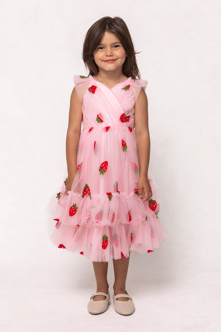 A young girl smiles in Ivy City Co’s Mini Marin Dress in Strawberry Shortcake, featuring pink tulle, strawberry embroidery, and ruffled layers, paired with white textured shoes against a plain white background.