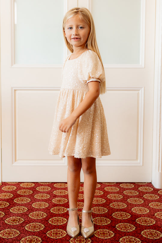 little girl in a cream sequin dress standing against a white wall