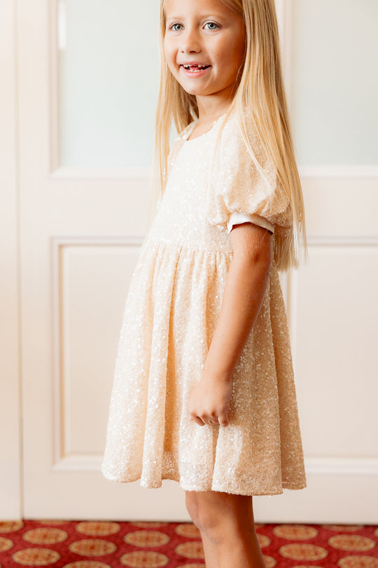 photo of a little girl in a cream sequin dress standing against a white wall