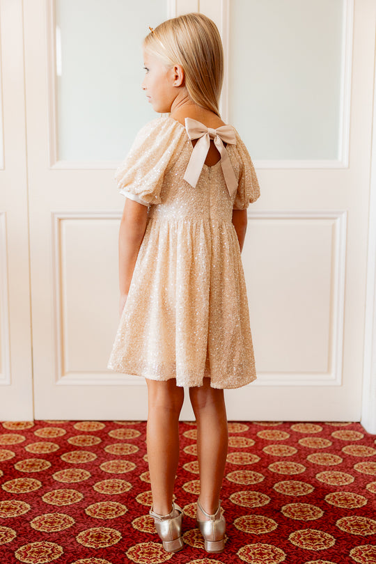 the back of a little girl in a cream sequin dress standing against a white wall