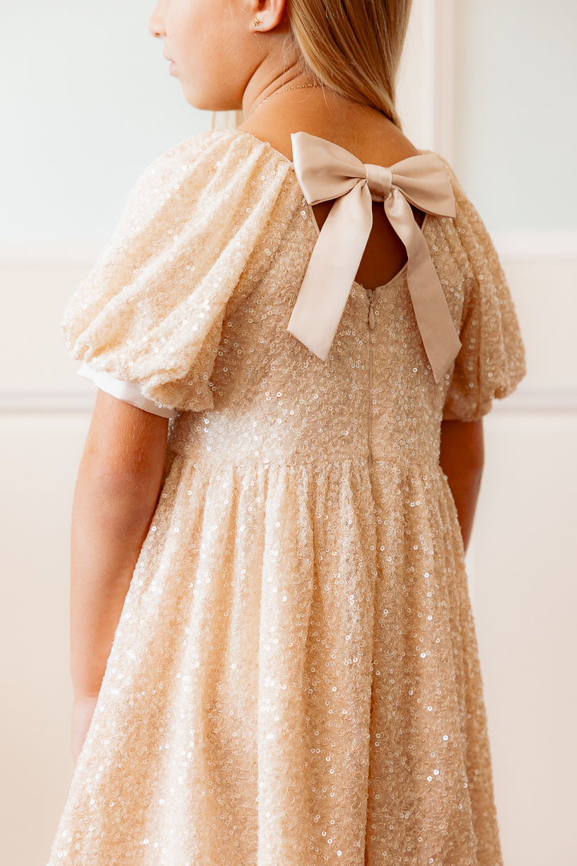 close up of the back of a little girl in a cream sequin dress standing against a white wall