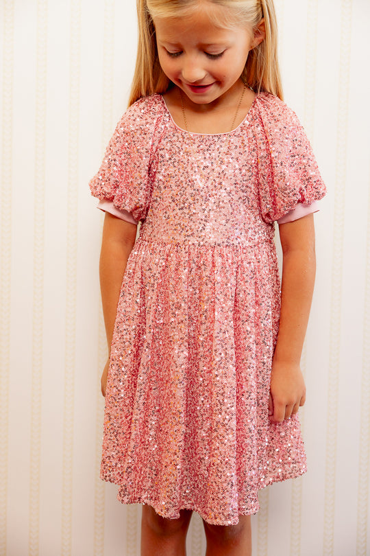 Close up of a little girl in a Pink sequin dress against a white wall standing on a red carpet