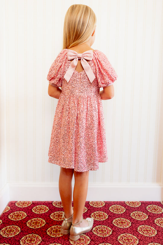 Little girl in a Pink sequin dress against a white wall standing on a red carpet with her back to the audience