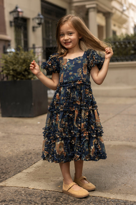 A young girl stands on a city sidewalk, smiling and holding her hair, wearing the Ivy City Co Mini Marseille Dress in Sunflower Twilight—a cheerful blue floral holiday dress—surrounded by buildings and plants, capturing the joyful spirit of summer evenings.