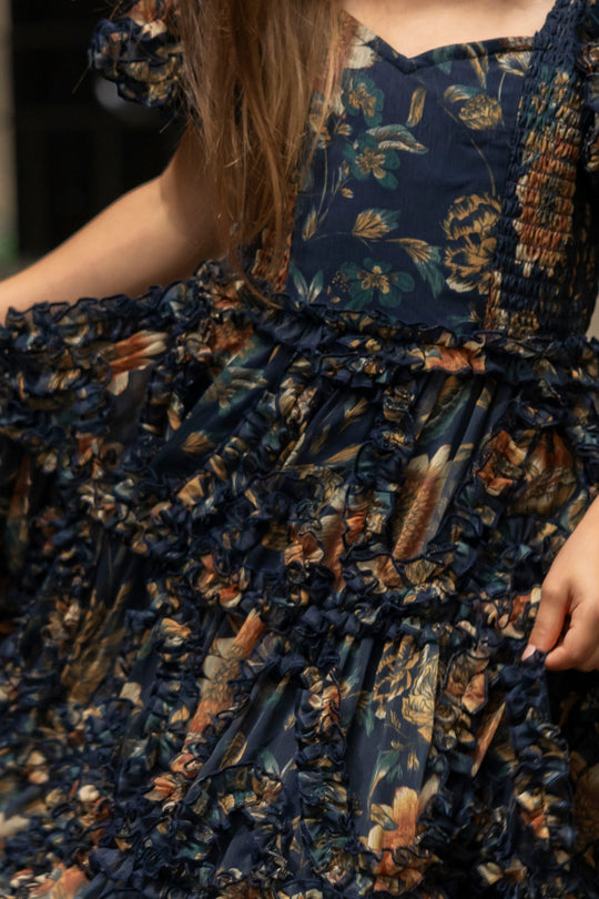 A child wearing the Ivy City Co Mini Marseille Dress in Sunflower Twilight with ruffles, holding out the skirt; only the lower face, torso, and part of the arms are visible—this charming girls holiday dress is perfect for special occasions.