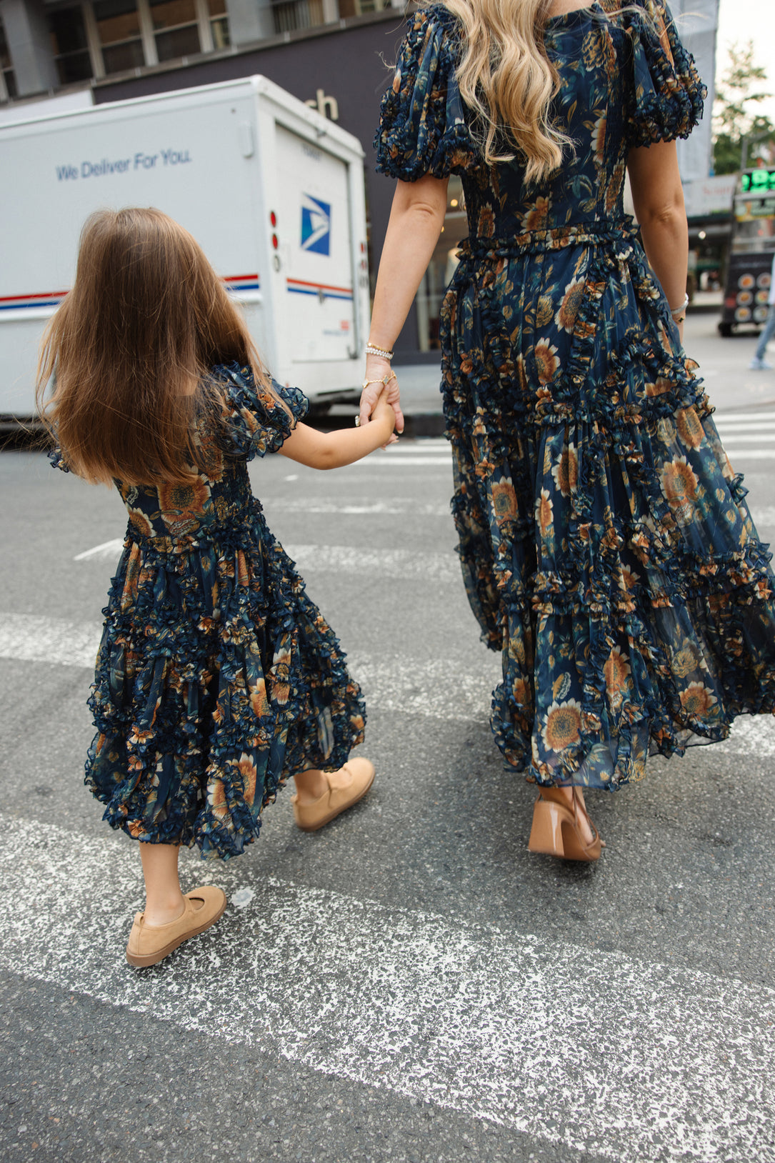 A woman and a young girl, both wearing matching Ivy City Co Mini Marseille Dresses in Sunflower Twilight with tan shoes, hold hands while crossing a city street. A USPS delivery truck is in the background, capturing a perfect mother-daughter moment in their stylish girls holiday dresses.