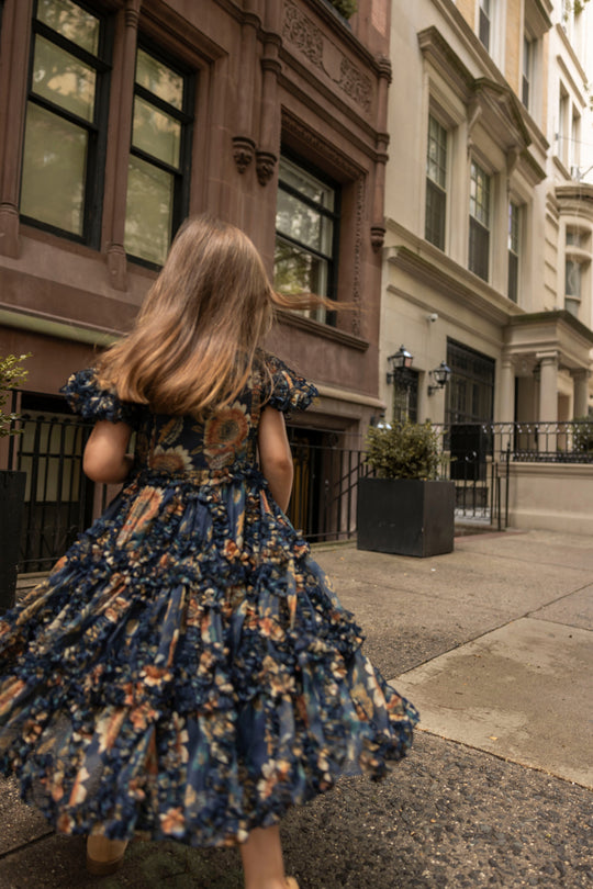 A young girl with long hair strolls along a city sidewalk lined with brownstone buildings, her Ivy City Co Mini Marseille Dress in Sunflower Twilight blooming vibrantly in the evening light.