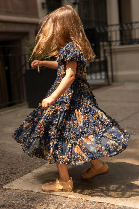 A young girl twirls with delight on a city sidewalk at sunflower twilight, her long hair swirling and her tan shoes peeking out beneath the Ivy City Co Mini Marseille Dress in Sunflower Twilight.