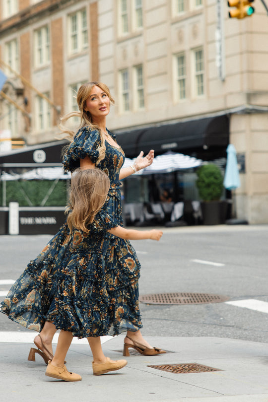 A woman and a young girl, both wearing matching floral dresses from Ivy City Co, walk across a city street. The woman looks upward while holding the girl’s hand, each dressed in coordinating holiday portrait ensembles—hers in the Mini Marseille Dress in Sunflower Twilight and the girl’s in an adorable girls midi dress. Buildings and outdoor dining areas are visible in the background.