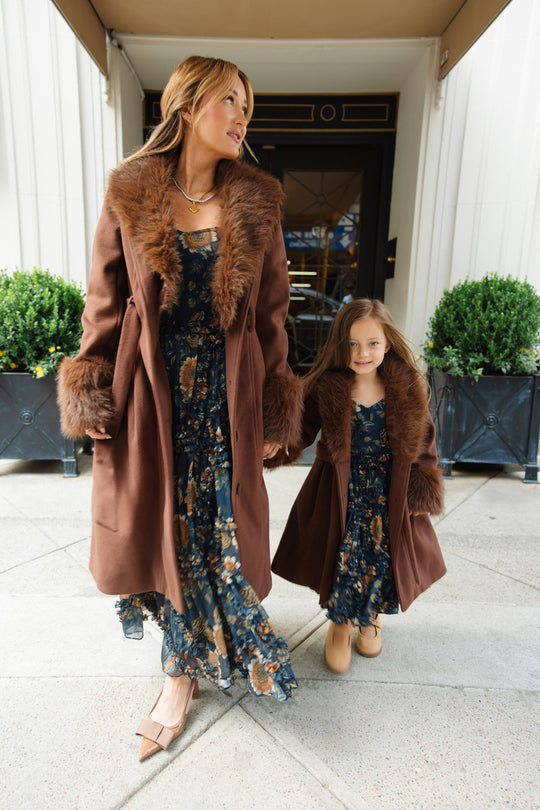 A woman and a young girl, likely mother and daughter, wear matching brown coats with fur trim over Ivy City Co’s Mini Marseille Dress in Sunflower Twilight—a blue floral girls’ midi dress—making for the perfect holiday portraits outfit as they walk together outside a building.